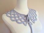 A flat lay view of a beautifully crocheted lace collar made with silky yarn. The handmade vintage-inspired collar features delicate shell openwork, a scalloped edge, and long integrated ties for adjusting the neckline.